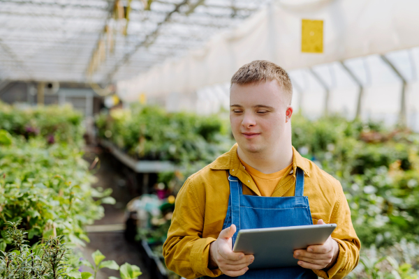 A young man with Down syndrome working in garden centre, holidng digital tablet and checking plants.