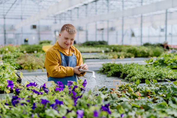 A happy young employee with Down syndrome working in garden centre, watering plants with a shower head and hose.