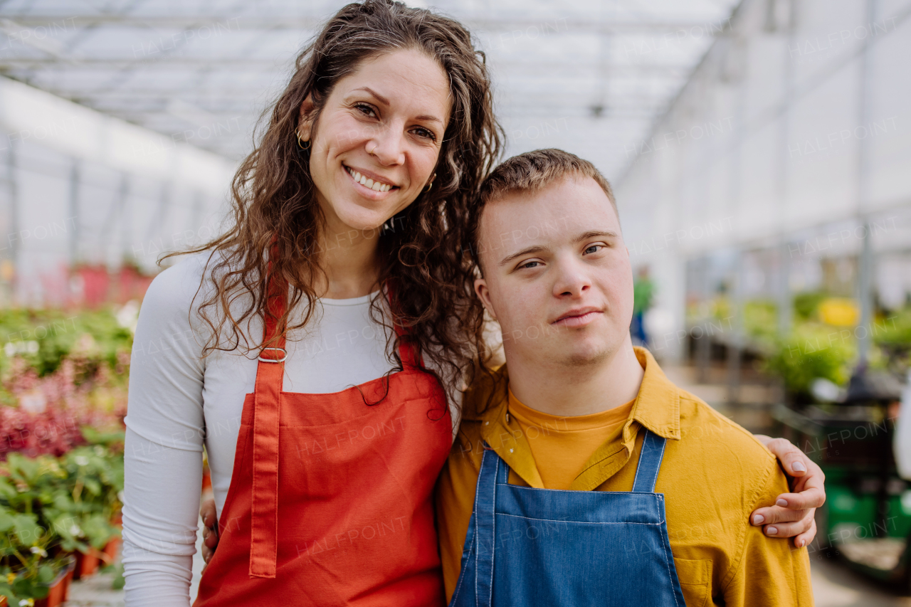 Experienced woman florist posing with young colleague with Down syndrome in garden centre, looking at camera.