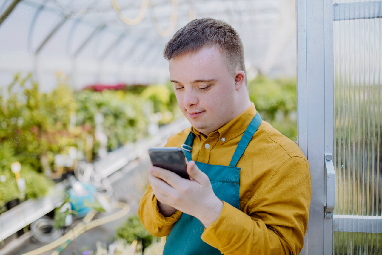 A young employee with Down syndrome working in garden centre, standing in door of greenhouse with cellphone.