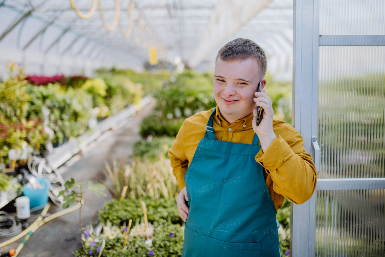 A young employee with Down syndrome working in garden centre, standing in door of greenhouse and calling on cellphone.