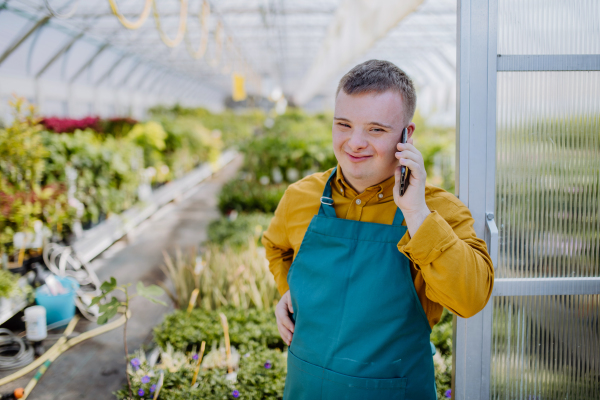 A young employee with Down syndrome working in garden centre, standing in door of greenhouse and calling on cellphone.