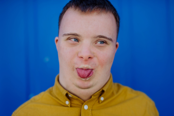 A close-up of happy young man with Down syndrome grimacing and sticking out tongue against blue background.