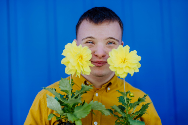 A happy young man with Down syndrome looking at camera and holding pot flowers against blue background.