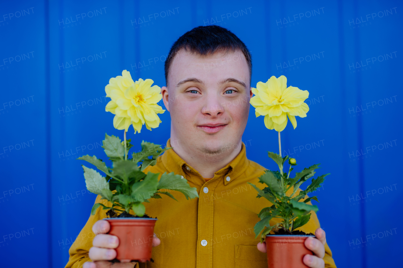 A happy young man with Down syndrome looking at camera and holding pot flowers against blue background.