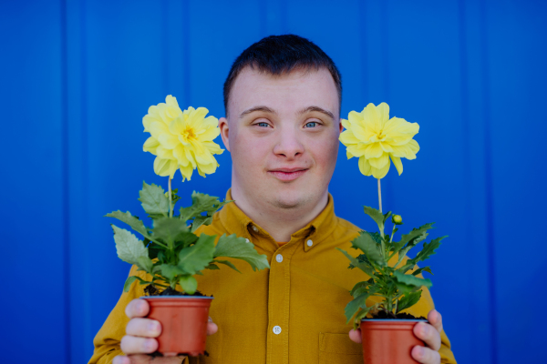 A happy young man with Down syndrome looking at camera and holding pot flowers against blue background.