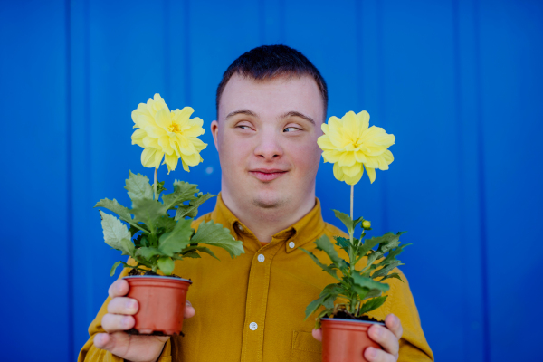 A happy young man with Down syndrome looking at camera and holding pot flowers against blue background.