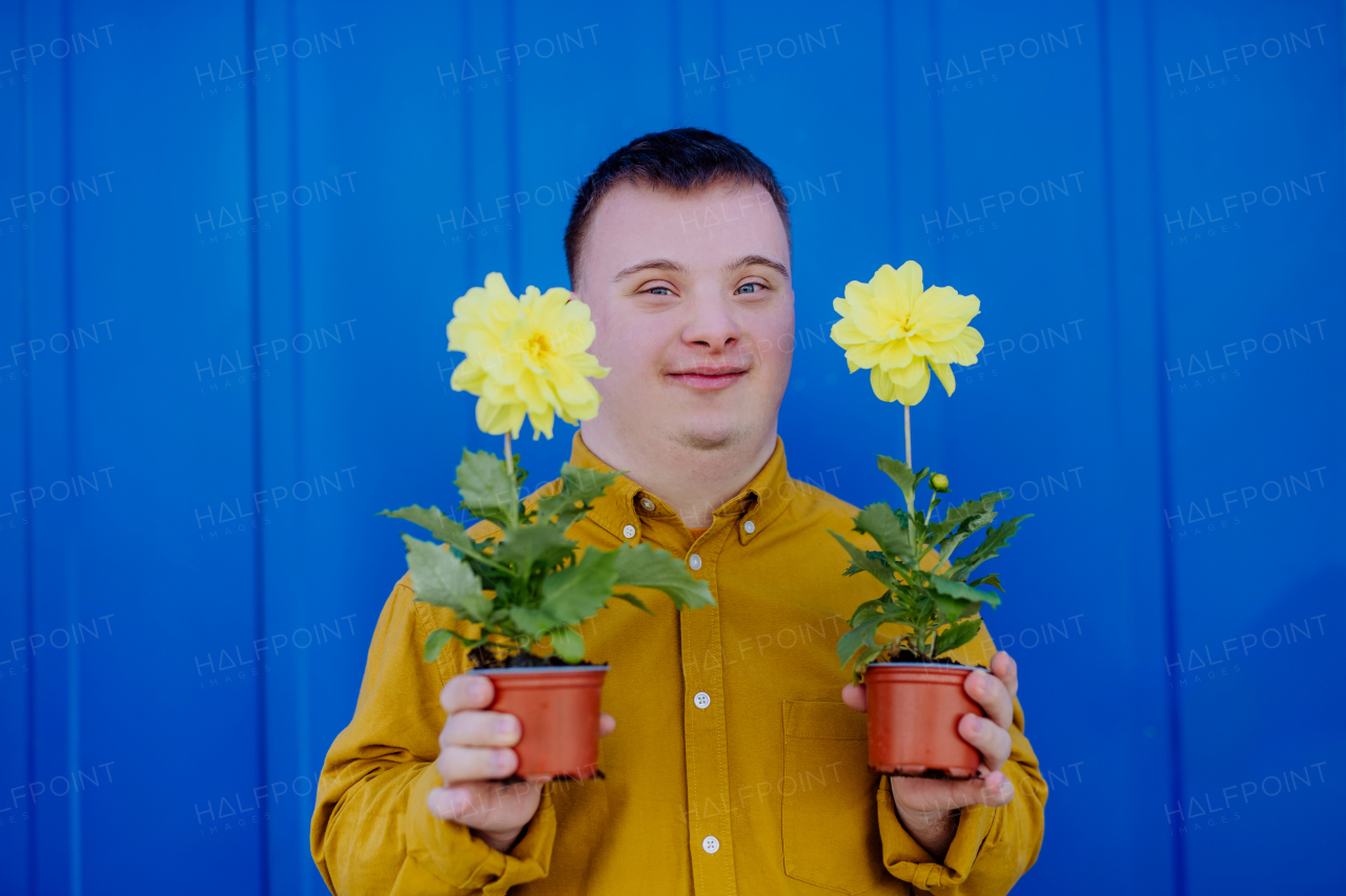 A happy young man with Down syndrome looking at camera and holding pot flowers against blue background.