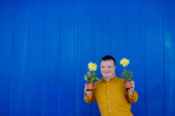 A happy young man with Down syndrome looking at camera and holding pot flowers against blue background.