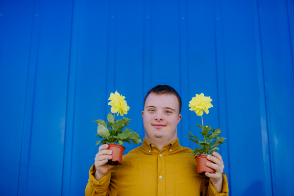 A happy young man with Down syndrome looking at camera and holding pot flowers against blue background.