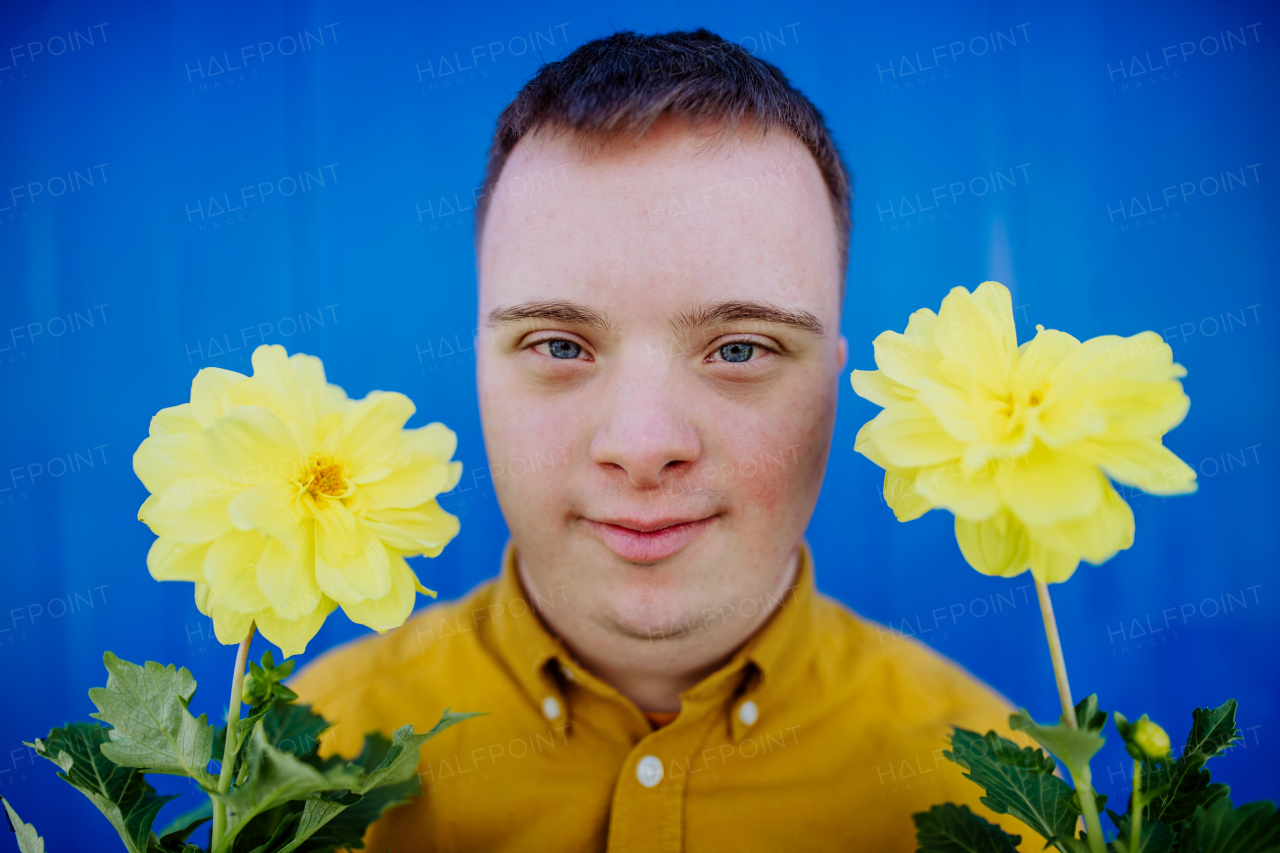 A happy young man with Down syndrome looking at camera and holding pot flowers against blue background.