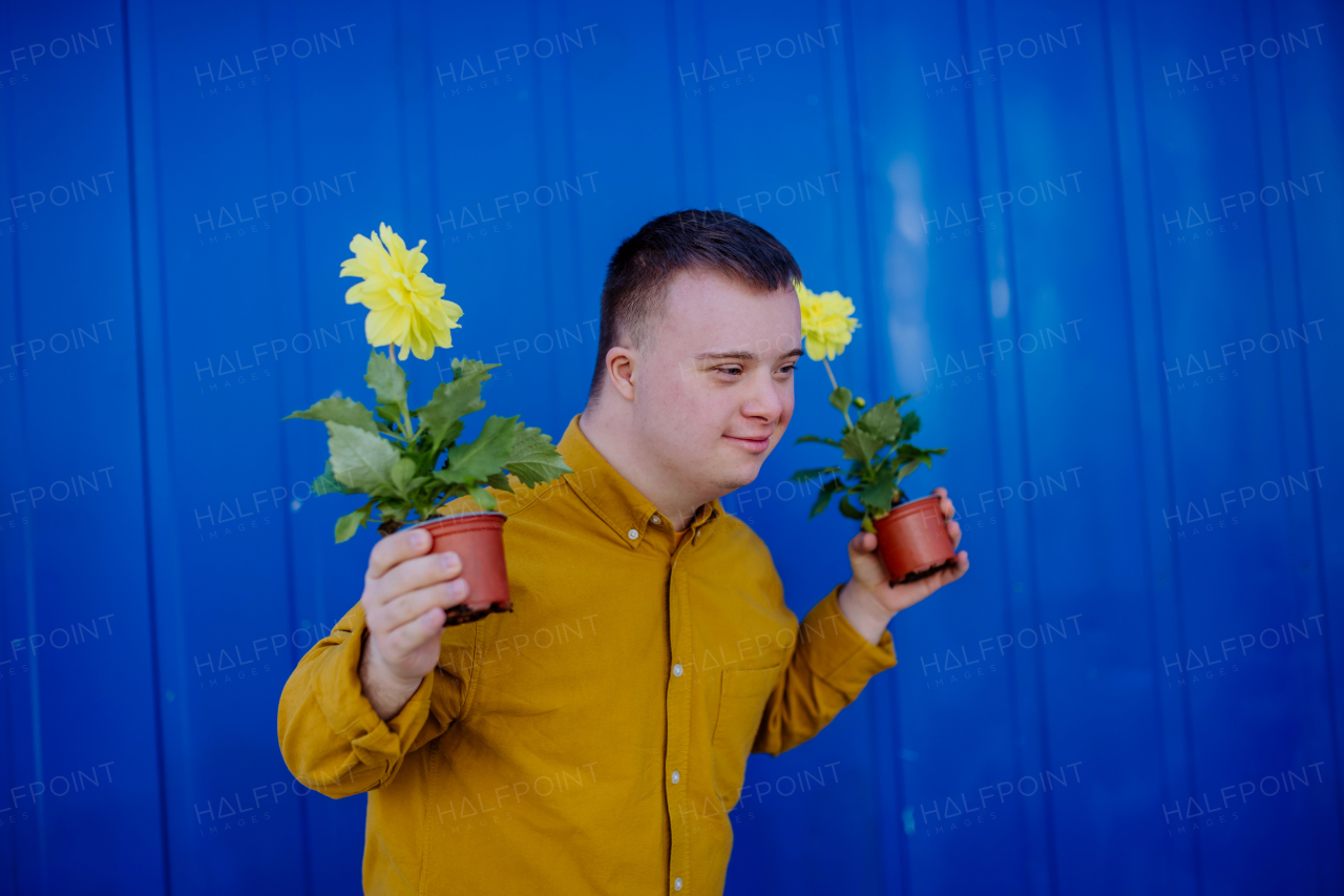 A happy young man with Down syndrome looking at camera and holding pot flowers against blue background.