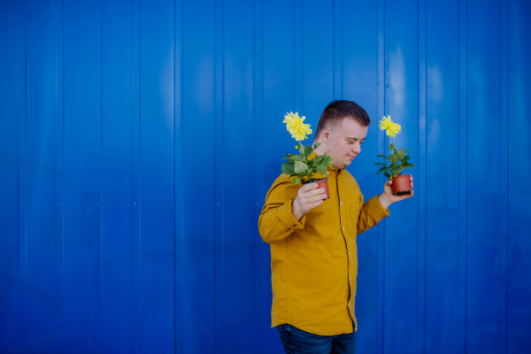 A happy young man with Down syndrome looking at camera and holding pot flowers against blue background.