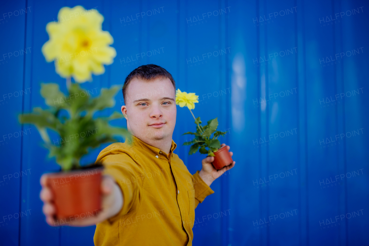 A happy young man with Down syndrome looking at camera and holding pot flowers against blue background.