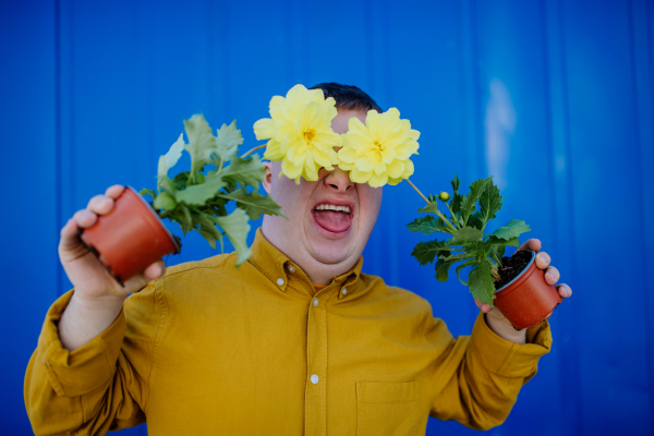 A happy young man with Down syndrome making funny grimace and holding pot flowers against blue background.