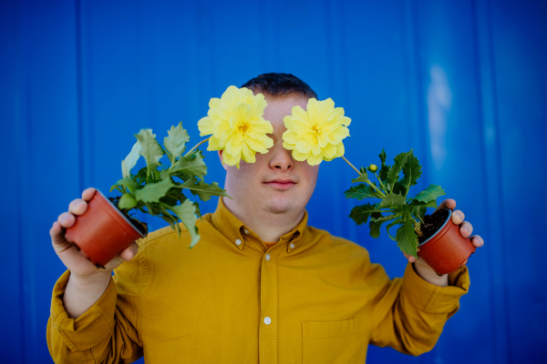 A happy young man with Down syndrome making funny grimace and holding pot flowers against blue background.