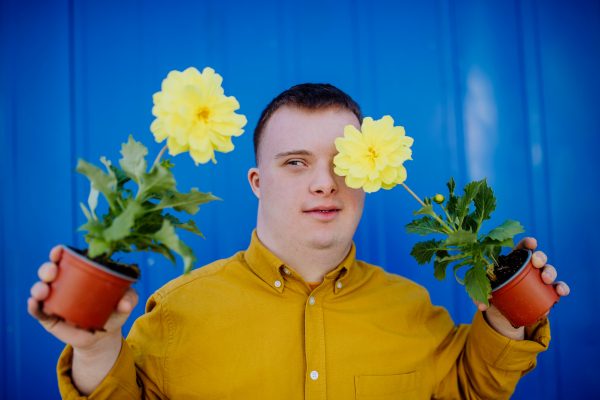 A happy young man with Down syndrome looking at camera and holding pot flowers against blue background.