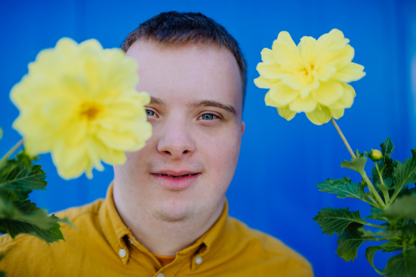 A happy young man with Down syndrome looking at camera and holding pot flowers against blue background.