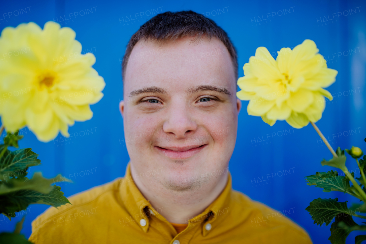 A happy young man with Down syndrome looking at camera and holding pot flowers against blue background.