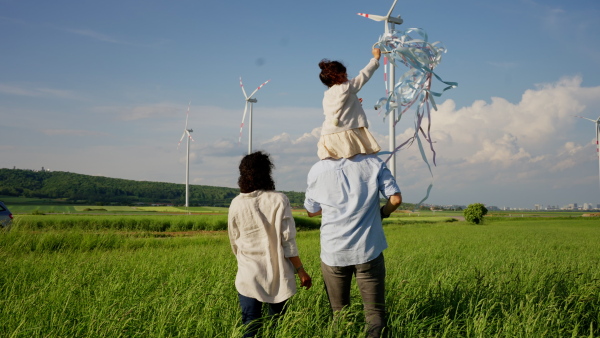 A rear view of multiracial family going for walk in field between wind turnines, concept of ecology and alternative energy