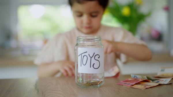 A cute little child girl putting coin into glass bottle at home. Kid saving money for toys.