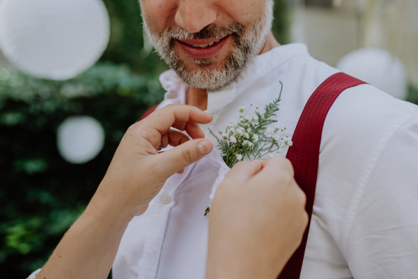 A mature groom recieving decoration on his shirt from bride at wedding reception outside in the backyard.