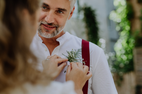 A mature groom recieving decoration on his shirt from bride at wedding reception outside in the backyard.