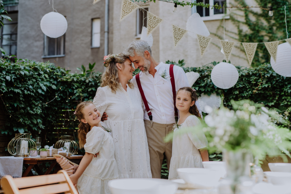 Mature bride and groom posing with their daughters at wedding reception outside in backyard.