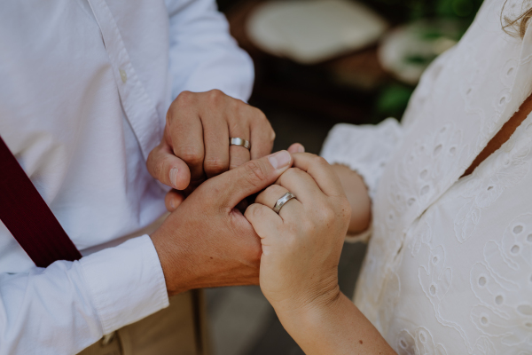 A close-up of bride and groom holding hands with wedding rings at reception outside in the backyard.