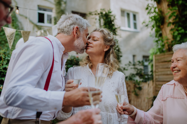 Mature bride and groom kissing at a wedding reception with their family, outside in the backyard.