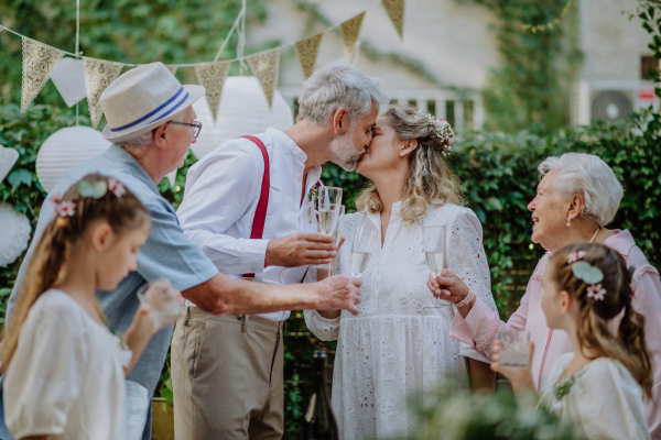 Mature bride and groom kissing at a wedding reception with their family, outside in the backyard.