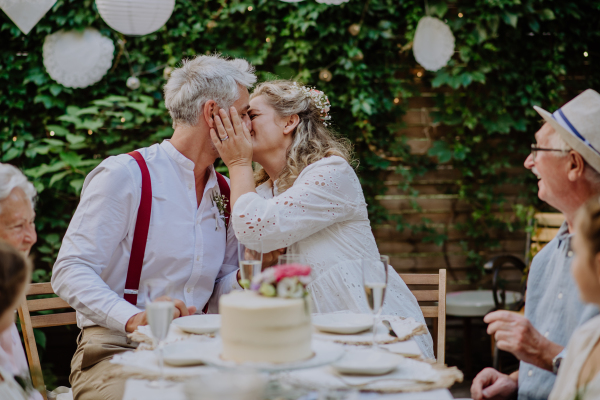 Mature bride and groom kissing at a wedding reception with their family, outside in the backyard.