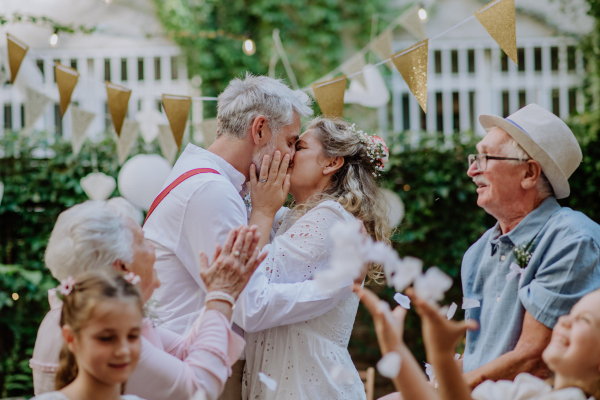 Mature bride and groom kissing at a wedding reception with their family, outside in the backyard.