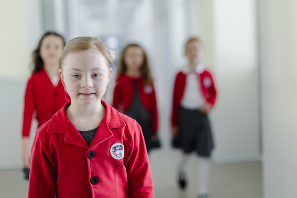 A happy schoolgrirl with Down syndrome in uniform walking in scool corridor with classmates behind her.