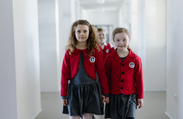 Happy schoolgrirl with Down syndrome in uniform walking in school corridor with her friend, holding each other hands.