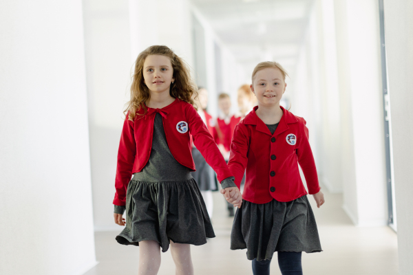 A happy schoolgrirl with Down syndrome in uniform holding hands with her classmate walking in scool corridor with classmates behind them.