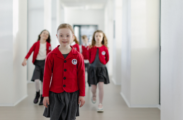 A happy schoolgrirl with Down syndrome in uniform walking in scool corridor with classmates behind her.