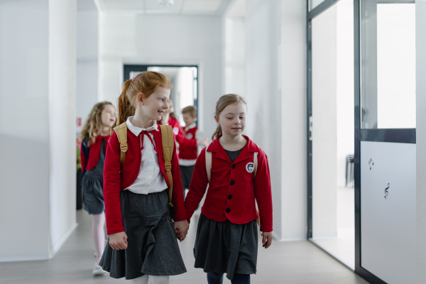 Happy schoolgrirl with Down syndrome in uniform walking in school corridor with her friend, holding each other hands.