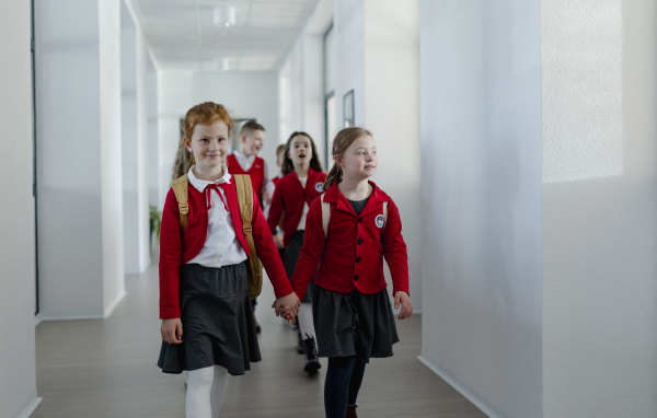 A happy schoolgrirl with Down syndrome in uniform holding hands with her classmate walking in scool corridor with classmates behind them.