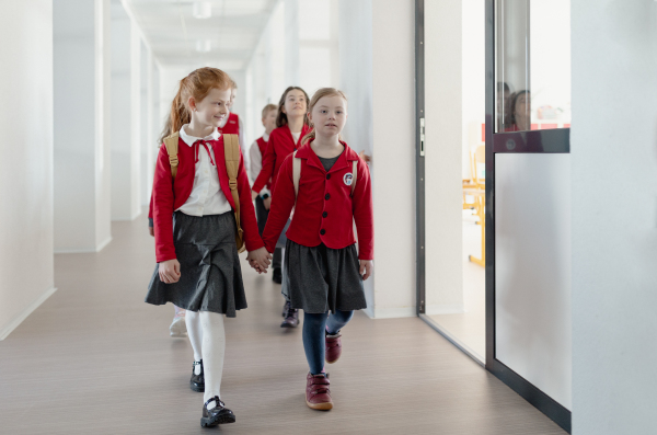 A happy schoolgrirl with Down syndrome in uniform holding hands with her classmate walking in scool corridor with classmates behind them.