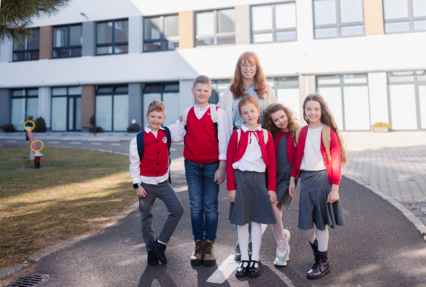 A portrait of happy schoolkids in uniforms with teacher at schoolyard, looking at camera.