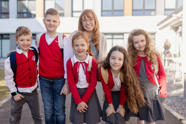 A portrait of happy schoolkids in uniforms with teacher at schoolyard, looking at camera.