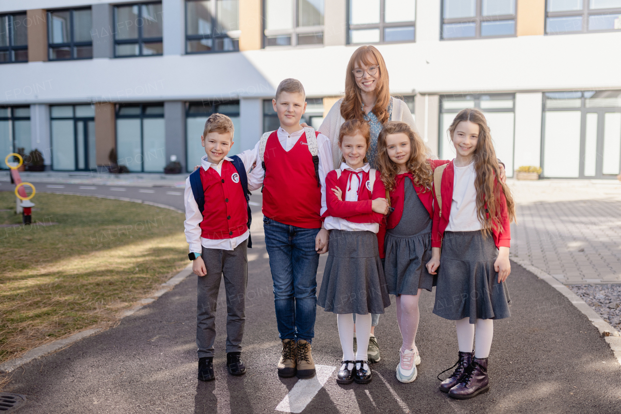 A portrait of happy schoolkids in uniforms with teacher at schoolyard, looking at camera.