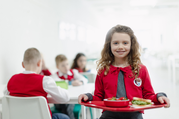 A happy schoolgrirl in uniform holding tray with lunch in school canteen.