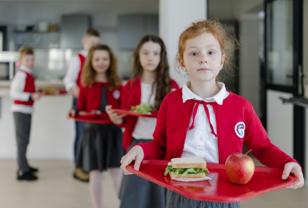 Happy schoolchildren in uniforms holding trays with lunch and standing in a queue in school canteen.