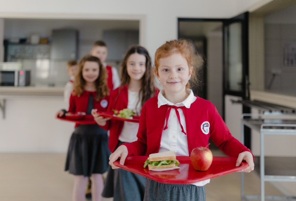 Happy schoolchildren in uniforms holding trays with lunch and standing in a queue in school canteen.