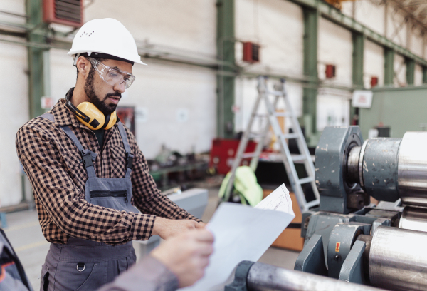 A heavy industry worker with safety headphones and hard hat in industrial factory holding blueprints