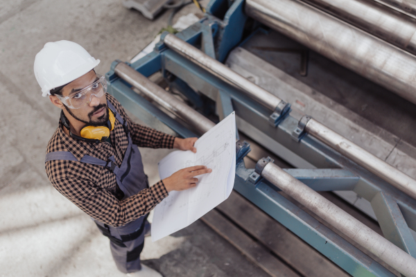 A high angle view of heavy industry worker with safety headphones and hard hat in industrial factory holding blueprints