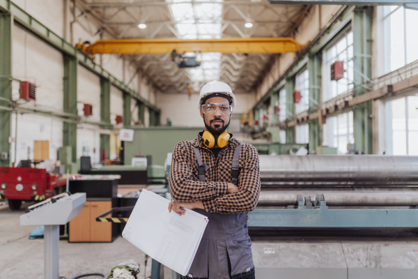 A heavy industry worker with safety headphones and hard hat in industrial factory holding blueprints