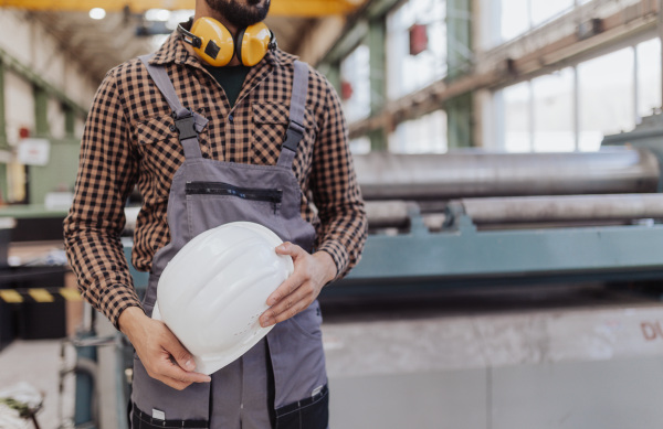 A midsection of heavy industry worker with safety headphones and hard hat in industrial factory